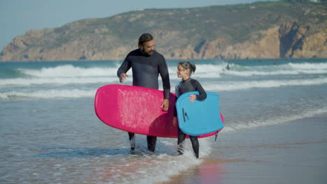 Long-Shot-Of-Surfer-With-Artificial-Leg-And-Daughter-Holding-Surfboard-And-Talking-While-Walking-On-The-Beach