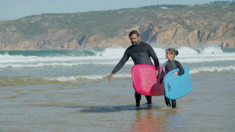 Surfer-With-Artificial-Leg-And-Daughter-Holding-Surfboard-And-Talking-While-Walking-On-The-Beach-1