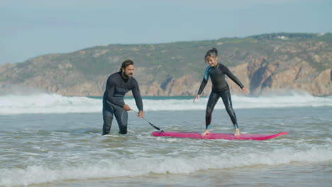 Long-Shot-Of-Happy-Girl-Standing-On-Surfboard-With-The-Help-Of-A-Coach