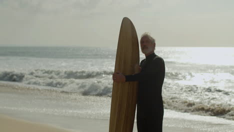 Senior-Man-With-Surfboard-Standing-On-The-Sandy-Beach-And-Dreaming