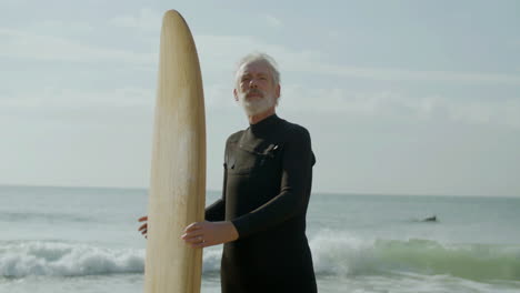 Porträt-Eines-älteren-Mannes-Mit-Surfbrett,-Der-Am-Sandstrand-Steht-Und-In-Die-Kamera-Schaut