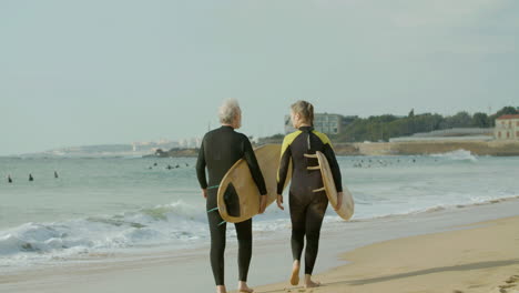 Back-View-Of-A-Senior-Couple-In-Wetsuit-Walking-Along-The-Beach-With-Surfboard