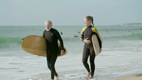 Senior-Couple-In-Wetsuit-Holding-Hands-And-Walking-Along-The-Beach-With-Surfboard