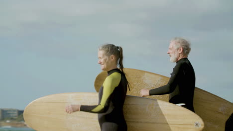 Mittlere-Aufnahme-Eines-älteren-Paares-Im-Neoprenanzug,-Das-Ein-Surfbrett-Hält-Und-Ins-Meer-Läuft