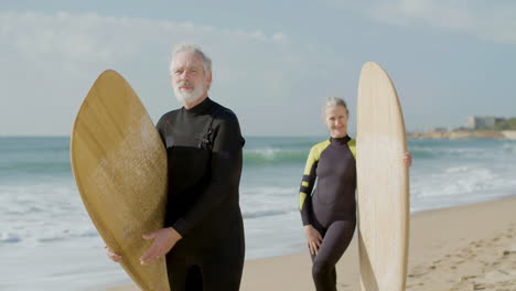 Front-View-Of-A-Senior-Couple-In-Wetsuit-With-Surfboard-Standing-On-The-Sandy-Beach-And-Looking-At-The-Camera