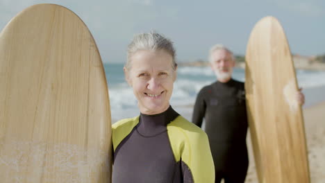 Front-View-Of-A-Happy-Senior-Woman-In-Wetsuit-With-Surfboard-Standing-On-The-Sandy-Beach-And-Looking-At-The-Camera-While-Her-Husband-Standing-Behind-In-The-Blurred-Background