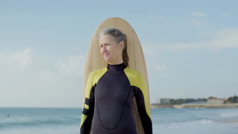 Front-View-Of-A-Cheerful-Senior-Woman-Standing-On-Seashore-With-Surfboard-And-Looking-At-The-Camera