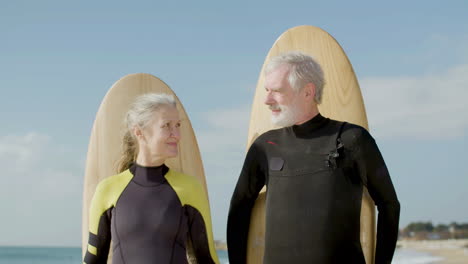 Vorderansicht-Eines-älteren-Paares-Mit-Surfbrett-Hinter-Dem-Rücken,-Das-Am-Meeresufer-Steht-Und-In-Die-Kamera-Schaut