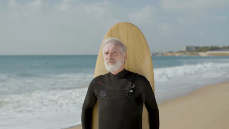 Front-View-Of-A-Smiling-Senior-Man-Standing-On-Seashore-With-Surfboard-And-Looking-At-The-Camera