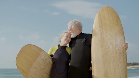 Front-View-Of-A-Senior-Couple-In-Wetsuit-With-Surfboard-Standing-On-The-Sandy-Beach,-Looking-At-The-Camera-And-Kissing