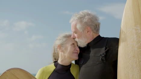 Close-Up-Of-A-Senior-Couple-In-Wetsuit-With-Surfboard-Standing-On-The-Sandy-Beach,-Looking-At-The-Camera-And-Kissing