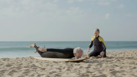 Seniorin-Erklärt-Ihrem-Mann,-Wie-Man-Surft,-Während-Er-Auf-Dem-Surfbrett-Am-Sandstrand-Liegt
