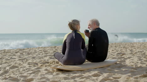 Long-Shot-Of-A-Senior-Couple-In-Wetsuit-Drinking-Tea-On-Beach-After-Surf-Training