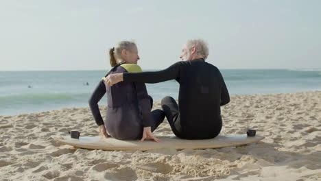 Back-View-Of-A-Senior-Couple-In-Wetsuit-Embracing-While-Sitting-On-A-Surfboard-At-The-Beach
