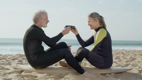 Senior-Couple-In-Wetsuit-Drinking-Tea-And-Talking-Together-While-Sitting-On-A-Surfboard-At-The-Beach