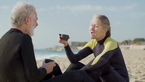 Senior-Couple-In-Wetsuit-Drinking-Tea-And-Talking-Together-While-Sitting-On-A-Surfboard-At-The-Beach-1
