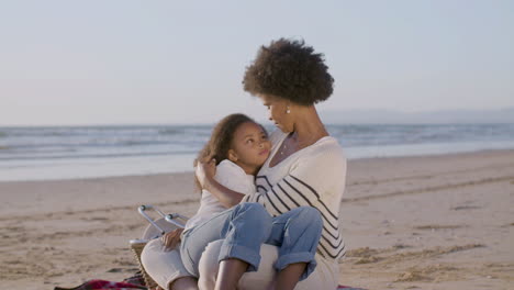 Happy-Woman-Holding-Her-Cute-Daughter-Lying-On-Her-Knees,-Stroking-Her-Curly-Hair-And-Talking-Together-During-A-Picnic-On-The-Beach
