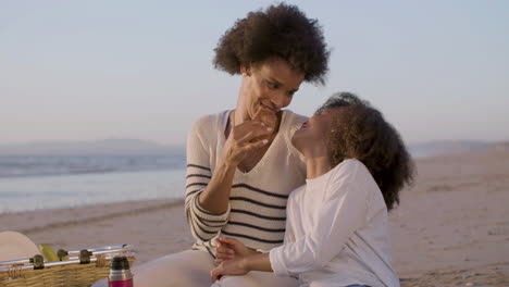 Liebevolle-Mutter-Und-Tochter-Teilen-Sich-Ein-Croissant-Und-Haben-Spaß-Bei-Einem-Picknick-Am-Strand