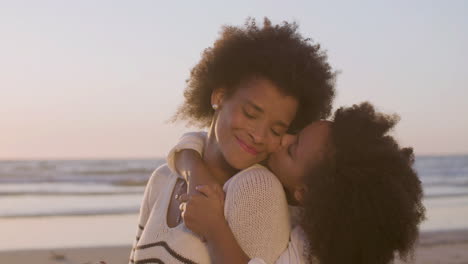 Cute-Girl-Hugging-Her-Mother-From-Behind-And-Kissing-Her-Cheek-Gently-While-Sitting-On-The-Beach-At-Sunset