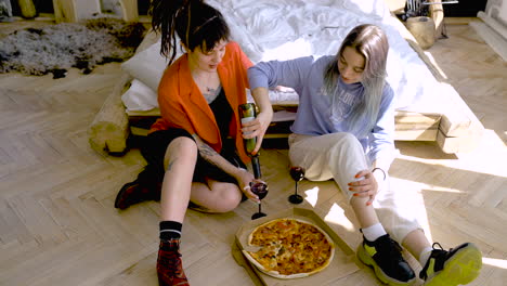 Zwei-Glückliche-Mädchen-Trinken-Wein-Und-Essen-Pizza,-Während-Sie-Zu-Hause-Auf-Dem-Boden-Sitzen