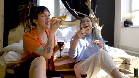 Two-Happy-Girls-Drinking-Wine-And-Sharing-Pizza-While-Sitting-On-The-Floor-At-Home