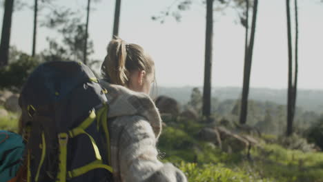 Two-Female-Friends-With-Backpacks-Hiking-Together-In-The-Forest-On-A-Sunny-Day