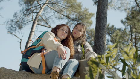 Two-Young-Female-Hikers-Sitting-On-A-Stone,-Hugging-And-Looking-At-The-Camera