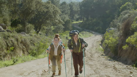 Front-View-Of-Two-Young-Female-Hikers-Walking-Up-Wide-Dirt-Road-With-Trekking-Poles-On-Bright-Autumn-Day
