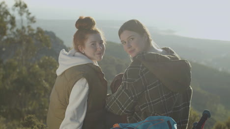 Two-Young-Female-Hikers-Sitting-At-Cliff-Edge-And-Talking,-Then-Turning-To-The-Camera-And-Smiling