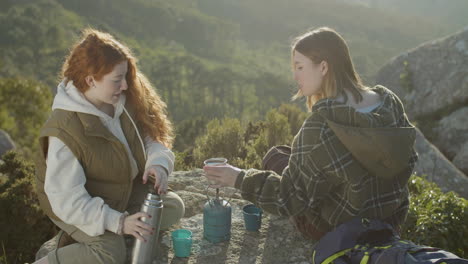 Two-Young-Female-Hikers-Drinking-Hot-Tea-From-Thermos-While-Sitting-At-Mountain-Cliff-On-A-Sunny-Autumn-Day