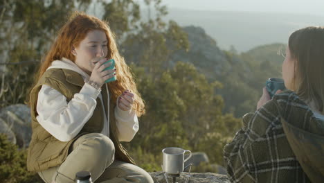 Two-Young-Female-Hikers-Drinking-Hot-Tea-From-Thermos-While-Sitting-At-Mountain-Cliff-On-A-Sunny-Autumn-Day-1