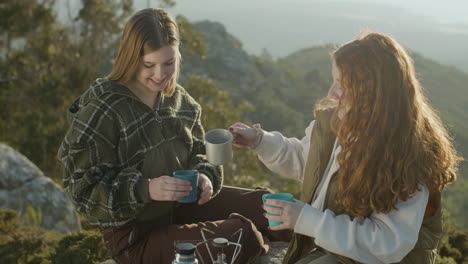 Two-Young-Female-Hikers-Drinking-Hot-Tea-From-Thermos-And-Talking-While-Sitting-At-Mountain-Cliff-On-A-Sunny-Autumn-Day