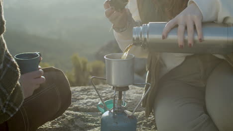 Close-Up-Shot-Of-An-Unrecognizable-Girl-Pouring-Hot-Tea-From-Thermos-Into-Mug-While-Sitting-At-Mountain-Top-And-Enjoying-Hiking-Trip-With-Friend