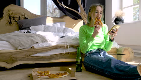 Junge-Blonde-Frau,-Die-Handy-Benutzt-Und-Pizza-Isst,-Während-Sie-Zu-Hause-Auf-Dem-Boden-Im-Schlafzimmer-Sitzt