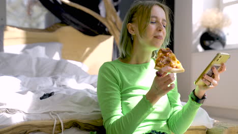 Young-Blonde-Woman-Watching-Something-Funny-On-Mobile-Phone-And-Eating-Pizza-While-Sitting-On-The-Floor-At-Home
