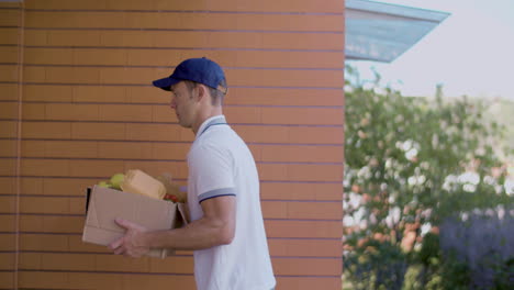 Side-View-Of-Male-Courier-With-Blue-Cap-Coming-To-Door-Of-Client-With-Box-Of-Groceries-And-Clipboard