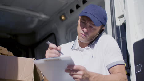 Focused-Courier-Calling-Customer-In-Van-And-Writing-On-Clipboard