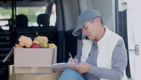 Delivery-Woman-Talking-With-Customer-On-Phone-In-A-Van-And-Writing-On-Clipboard-Next-To-Grocery-Box
