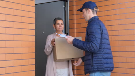 Woman-Standing-In-Doorway-And-Receiving-Parcel-From-Courier-And-Signing-Clipboard