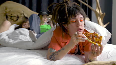 Young-Woman-With-Piercings-And-Tattoos-Eating-Pizza-While-Her-Friends-Are-Behind-Drinking-Wine-And-Relaxing