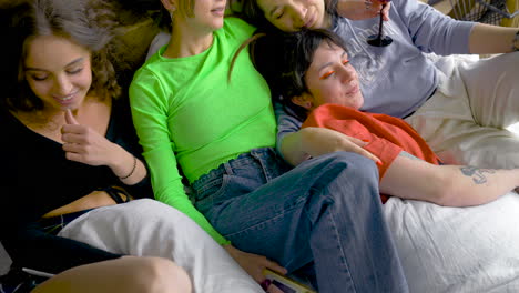 Four-Women-Laying-On-Bed-While-Talk,-Drink-And-Take-Pictures