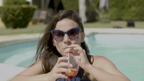 Front-View-Of-Happy-Woman-With-Blue-Swimsuit-And-Sunglasses,-Drinking-Cocktail-In-Swimming-Pool