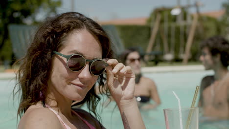Close-Up-Of-Cute-Girl-Standing-In-Swimming-Pool-With-Cocktail,-Playing-With-Her-Sunglasses-While-Her-Friends-Have-Fun-At-The-Middle-Of-The-Pool