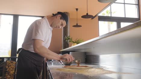 Vista-Lateral-De-Un-Chef-Esparciendo-Salsa-Sobre-Masa-De-Pizza-En-La-Encimera-De-La-Cocina-De-Un-Restaurante
