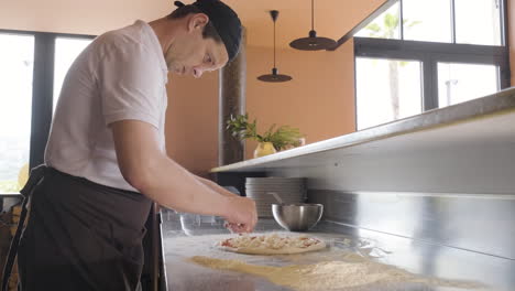 Side-View-Of-A-Chef-Spreading-Cheese-On-Pizza-Dough-On-A-Restaurant-Kitchen-Countertop