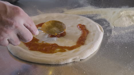 Vista-De-Cerca-De-Un-Chef-Esparciendo-Salsa-Sobre-Masa-De-Pizza-En-La-Encimera-De-La-Cocina-De-Un-Restaurante