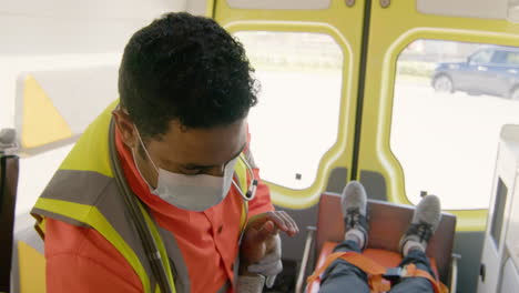 Paramedic-Wearing-Facial-Mask-Touching-The-Hand-And-Arm-Of-A-Patient-Lying-On-The-Stretcher-Inside-An-Ambulance
