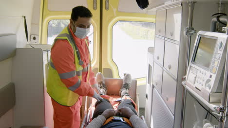 Paramedic-Wearing-Facial-Mask-Touching-The-Knee-Of-A-Patient-Lying-On-The-Stretcher-Inside-An-Ambulance