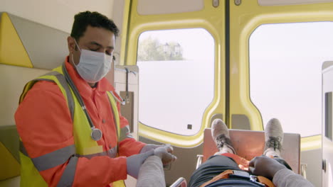 Paramedic-Wearing-Facial-Mask-Touching-The-Hand-Of-A-Patient-Lying-On-The-Stretcher-Inside-An-Ambulance
