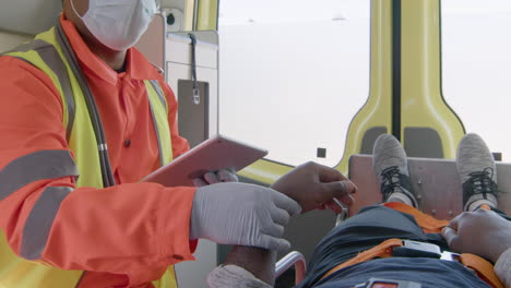 Male-Paramedic-With-Face-Mask-Using-Tablet-Computer-And-Taking-The-Pulse-Of-An-Injured-American-Patient-Who-Lying-On-The-Ambulance-Stretcher